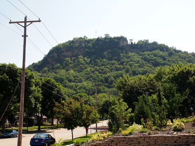 [Rock has been excavated at several levels of the bluff so some stone is visible amoung the leafy greenery on the hillside. Trees lining the street in town block some of the bluff from this angle.]